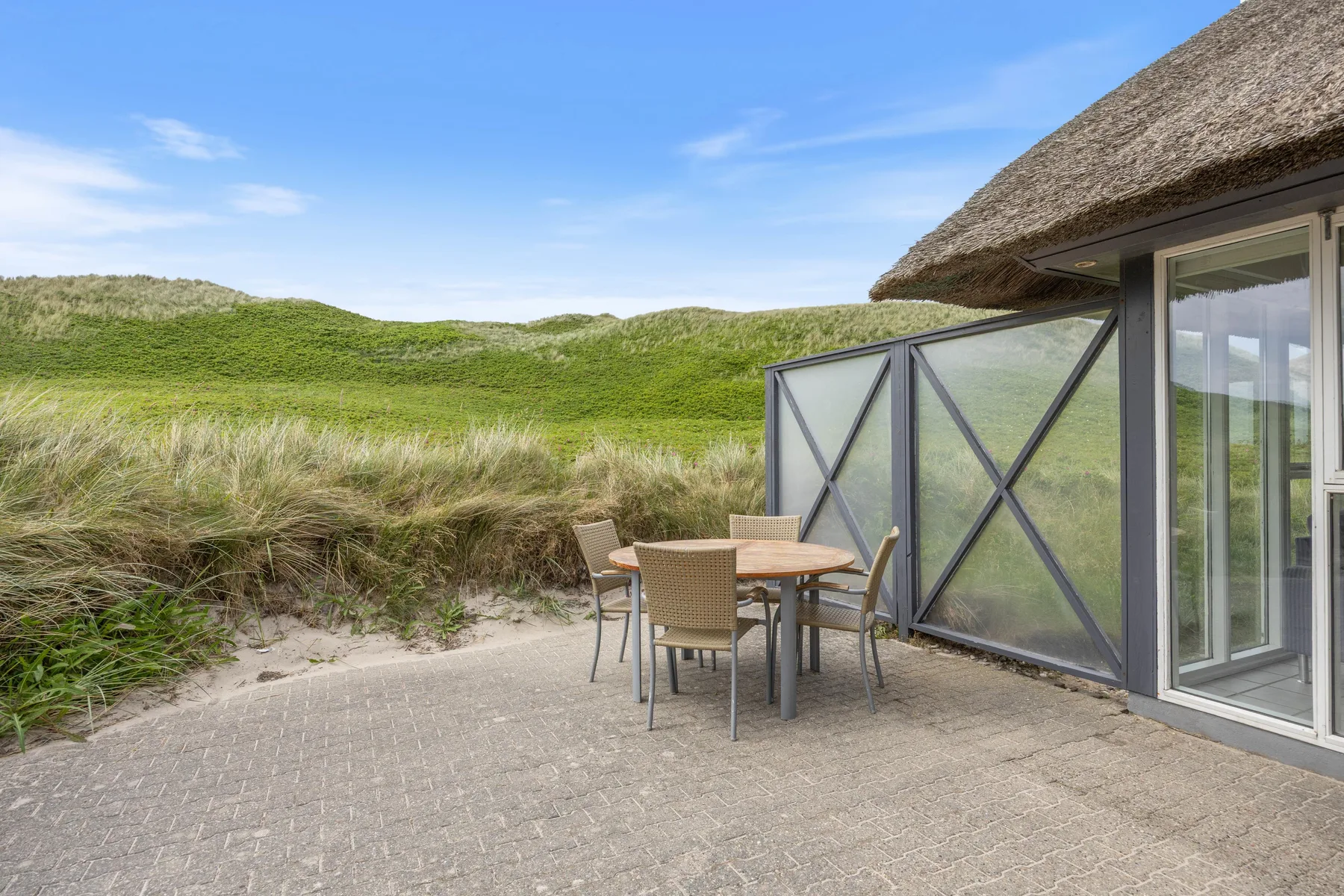 Un patio pavimentado junto a una casa, con asientos al aire libre y dunas verdes al fondo.