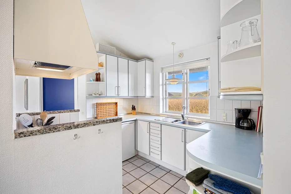 Una cocina luminosa con gabinetes blancos, suelo de baldosas y una ventana sobre el fregadero.
