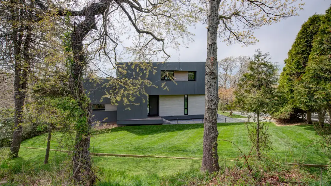 A modern dark-colored house stands beyond a green lawn and mature trees.