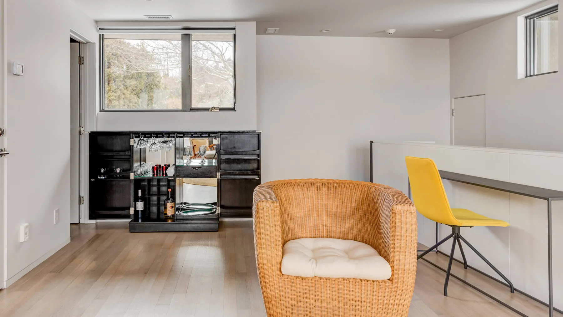 A bright living area with a woven armchair, yellow seat, and large windows.