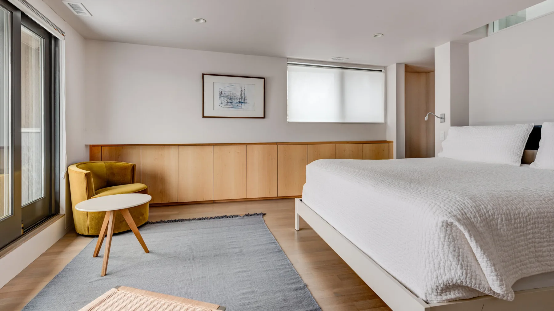 A bright bedroom with a bed, a small seating area, and large windows.