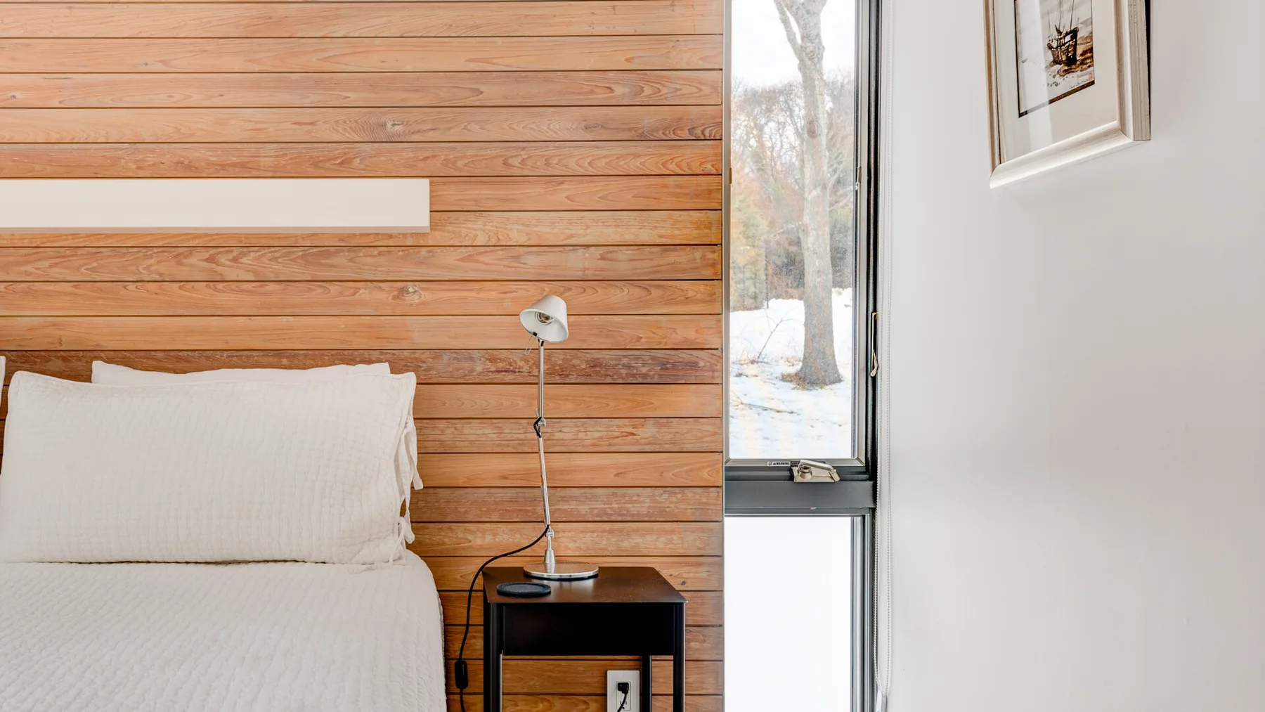 A bedroom with a bed beside a wooden wall and a large window.