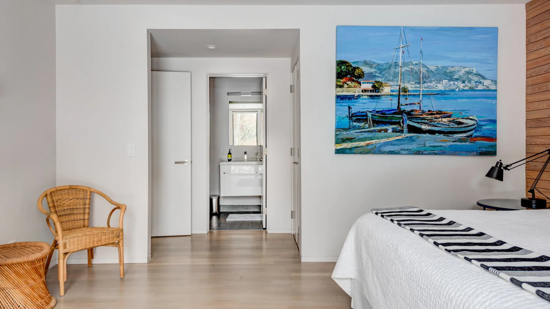 A bedroom with a bed, a woven chair, and a colorful painting on the wall.