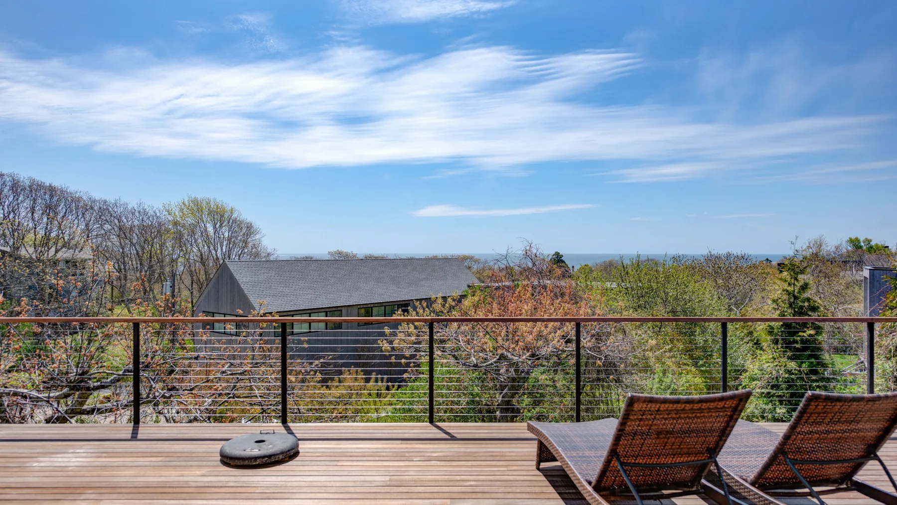 An outdoor deck with lounge seating overlooks leafy trees beneath a blue sky.