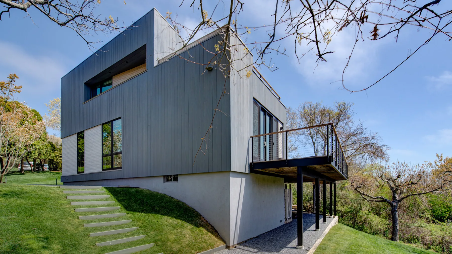 Una casa moderna se alza en una ladera cubierta de césped, con una terraza elevada y árboles alrededor.