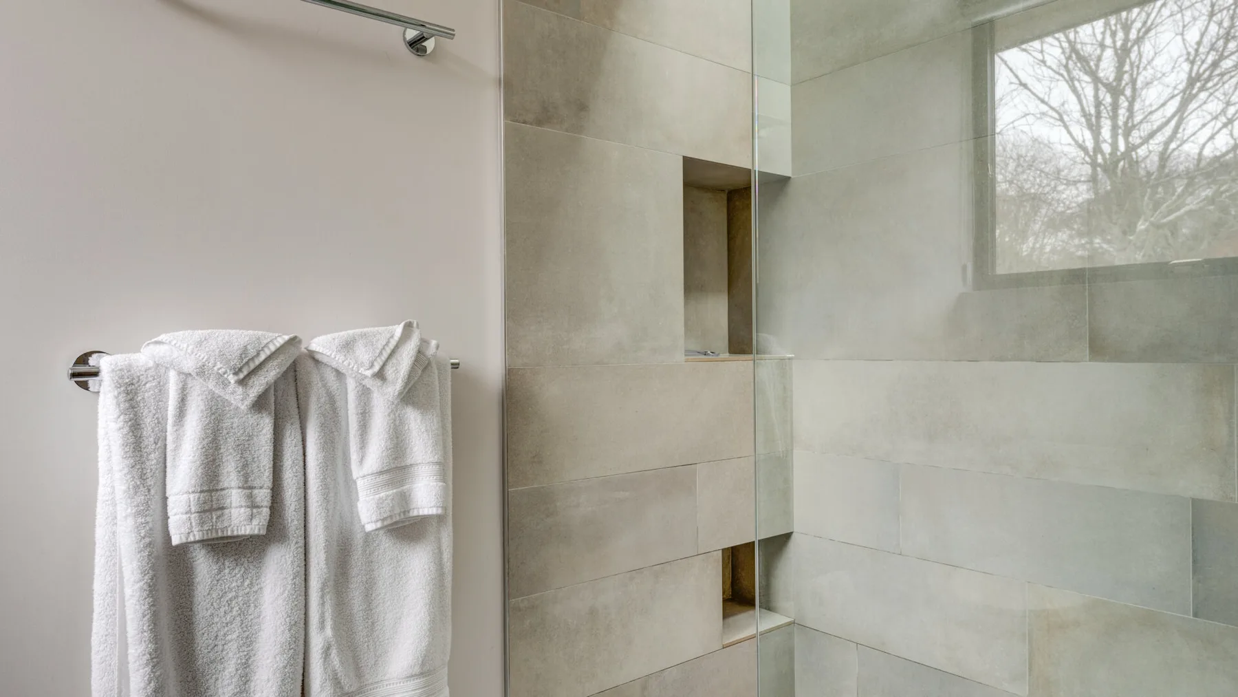 A bathroom shower with pale tiled walls and white towels hanging nearby.