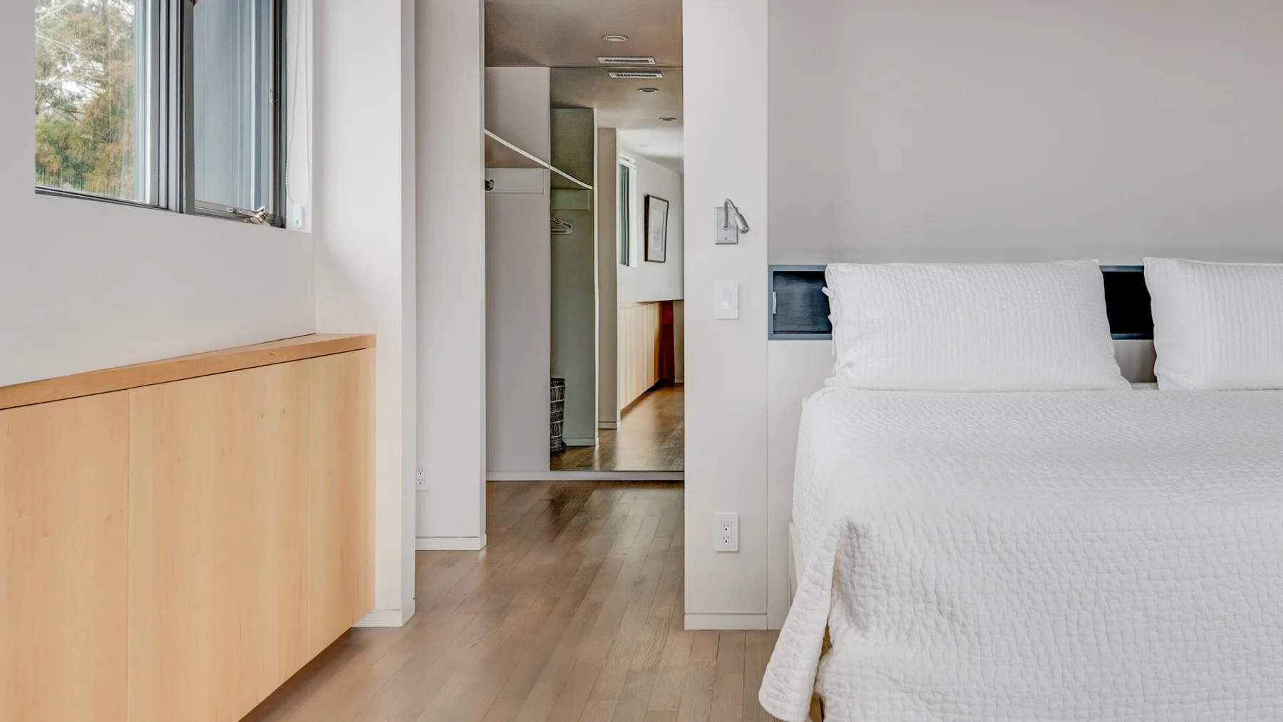 A bedroom with a bed beside a tall window and light wood flooring.