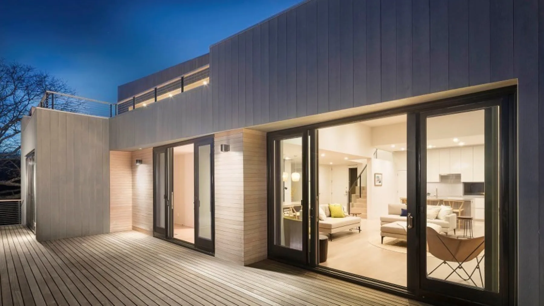 Modern house exterior with a broad deck and brightly lit interior visible through glass doors.