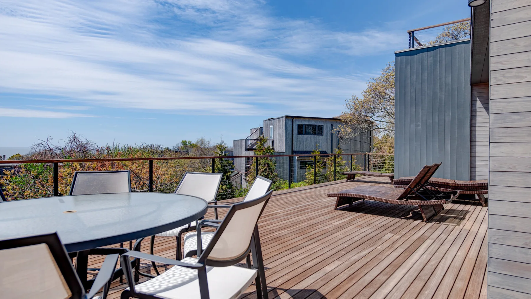A wooden outdoor deck has a dining table and lounge seating overlooking trees.