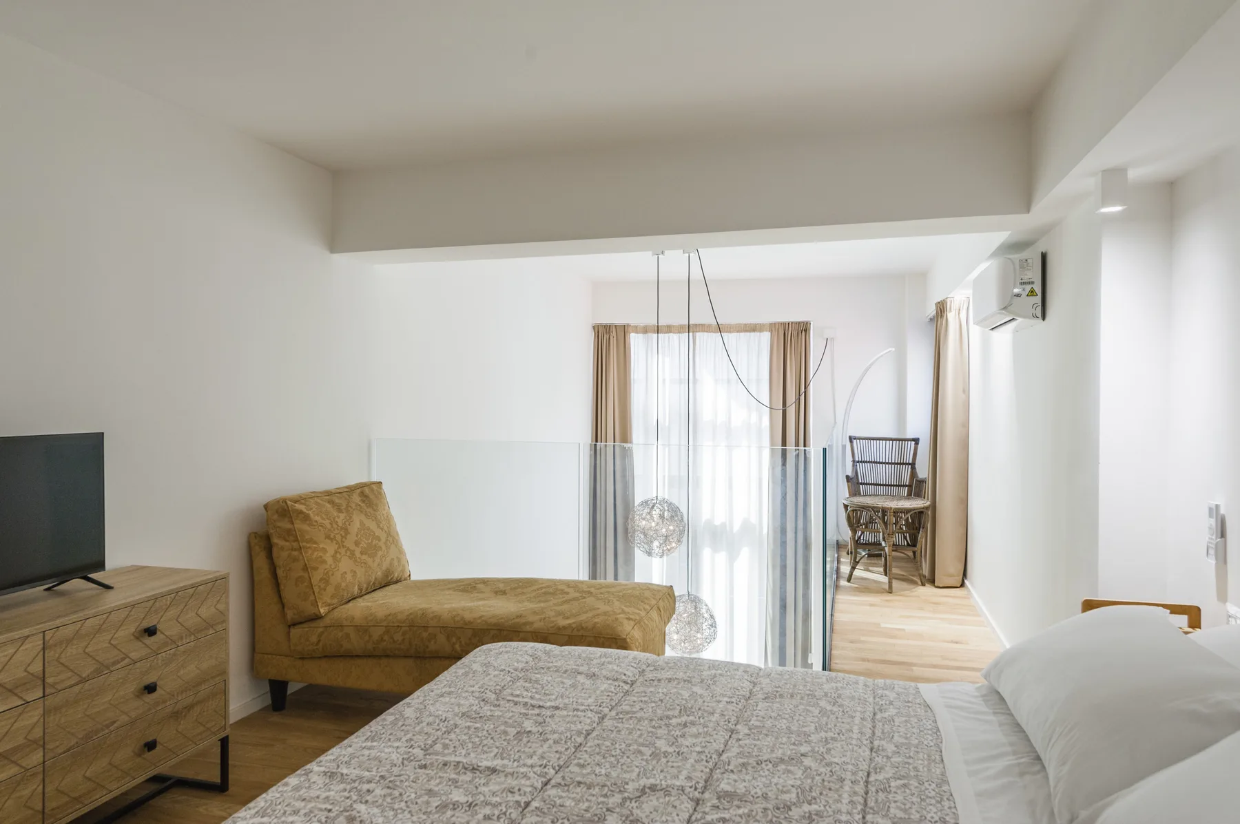Un dormitorio luminoso con cama, asientos y una mampara de vidrio junto a la ventana.