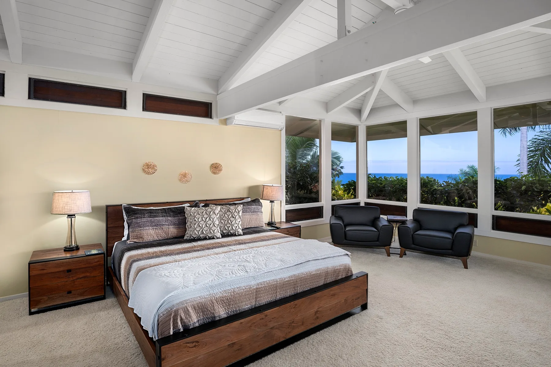 Un dormitorio con una cama grande y asientos junto a amplias ventanas con vistas a vegetación y agua.