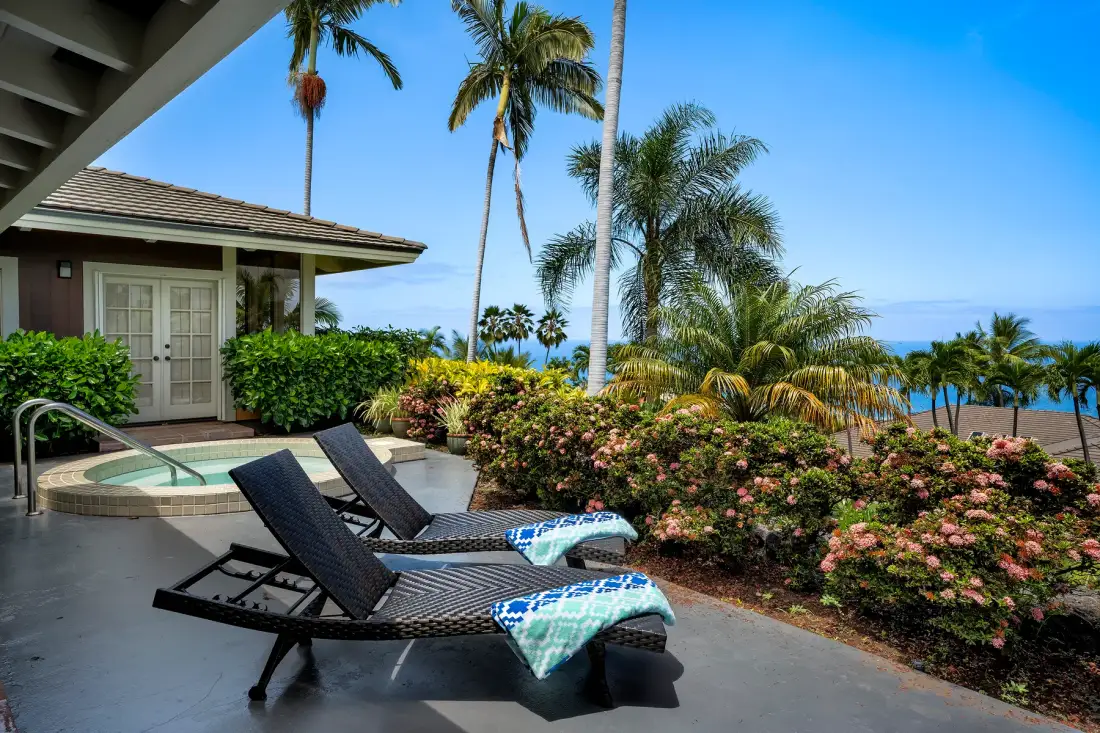 Un patio junto a la piscina con asientos reclinables, plantas tropicales y vista al mar.
