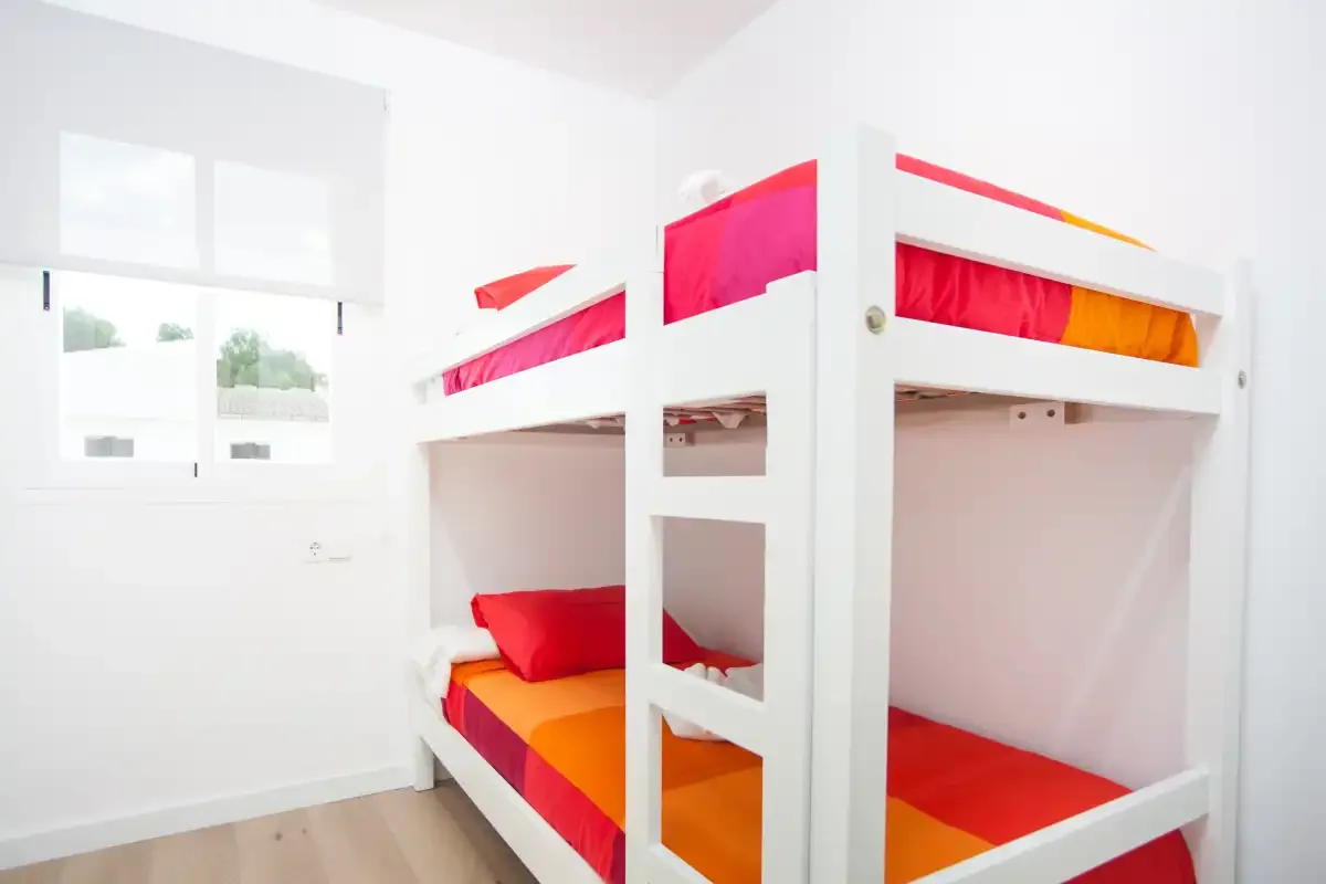 Un dormitorio luminoso con literas blancas y ropa de cama colorida junto a una ventana.