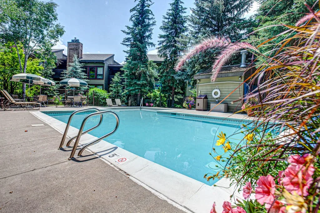 An outdoor swimming pool is surrounded by trees, patio seating, and flowering plants.