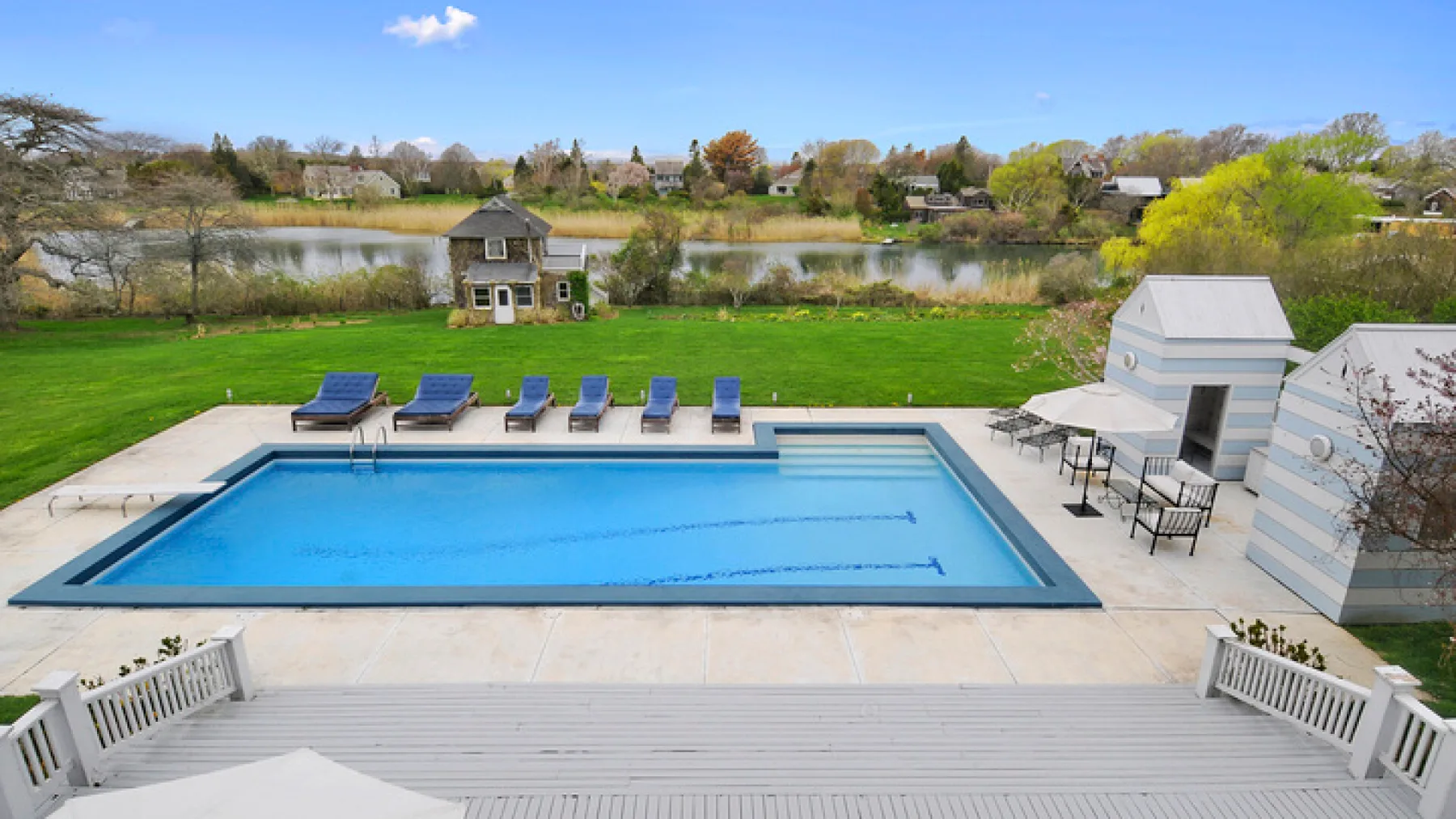 Una piscina al aire libre junto a una amplia terraza, con un estanque y árboles al fondo.