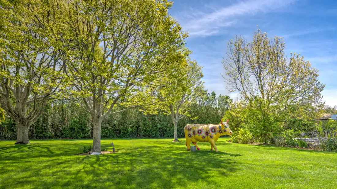 Un jardín con césped, árboles frondosos y una colorida escultura de vaca.