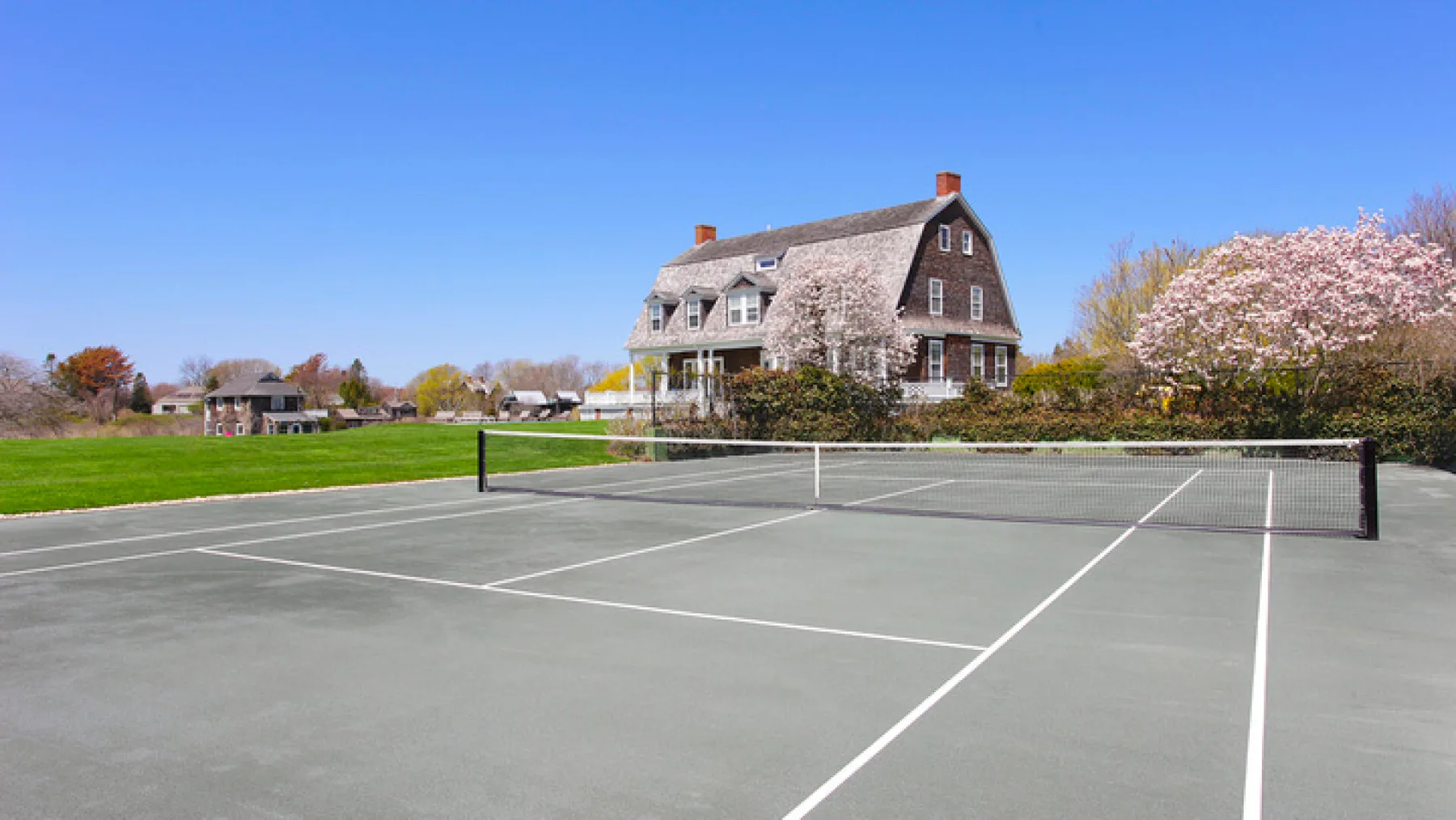 Una cancha de tenis está frente a una casa grande y árboles en flor.