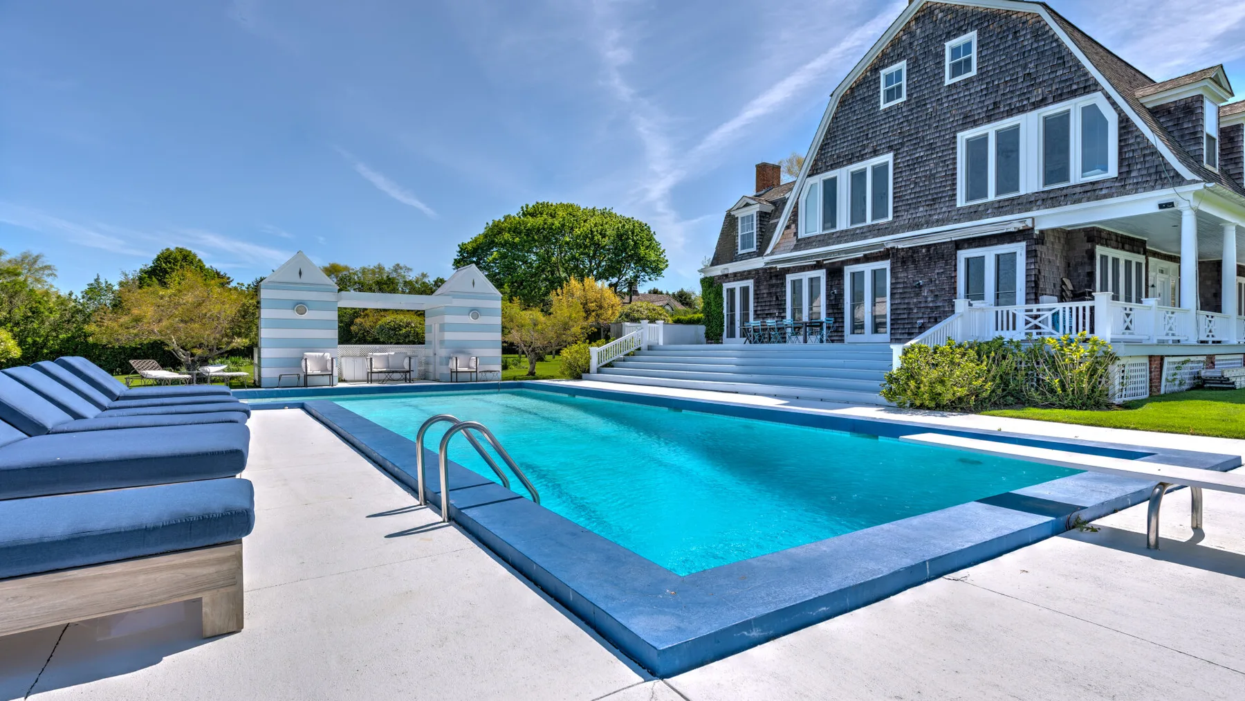 Una piscina exterior junto a una casa grande, con asientos de descanso en el patio.