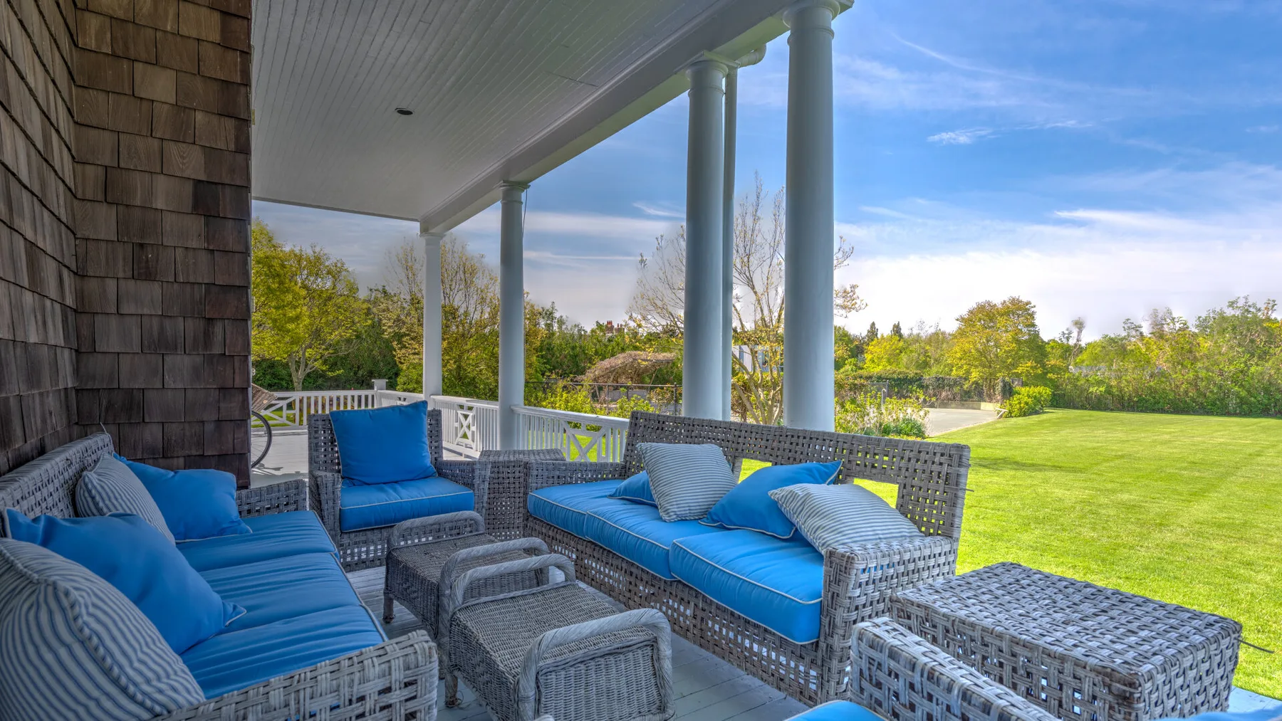 Un patio cubierto tiene asientos con cojines azules junto a un amplio césped verde.