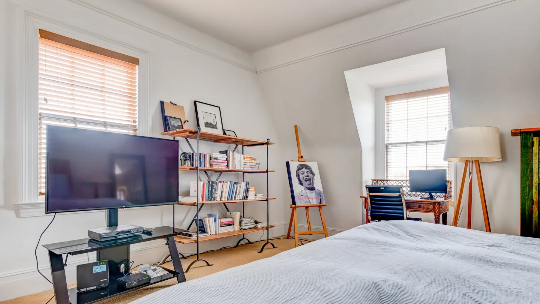 Un dormitorio con cama, televisor, estanterías y un escritorio junto a la ventana.