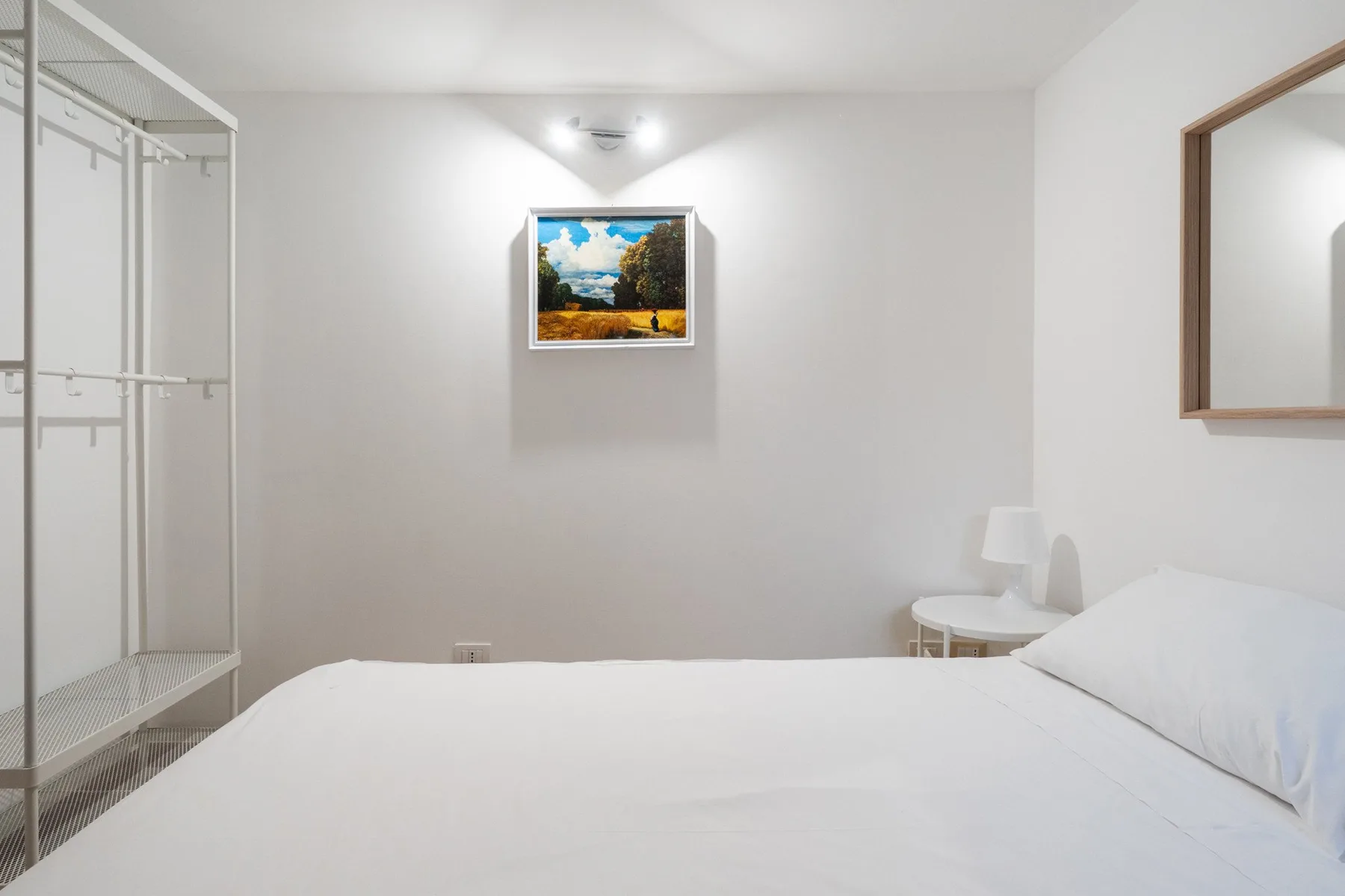 Un dormitorio luminoso con una cama blanca y un paisaje enmarcado.