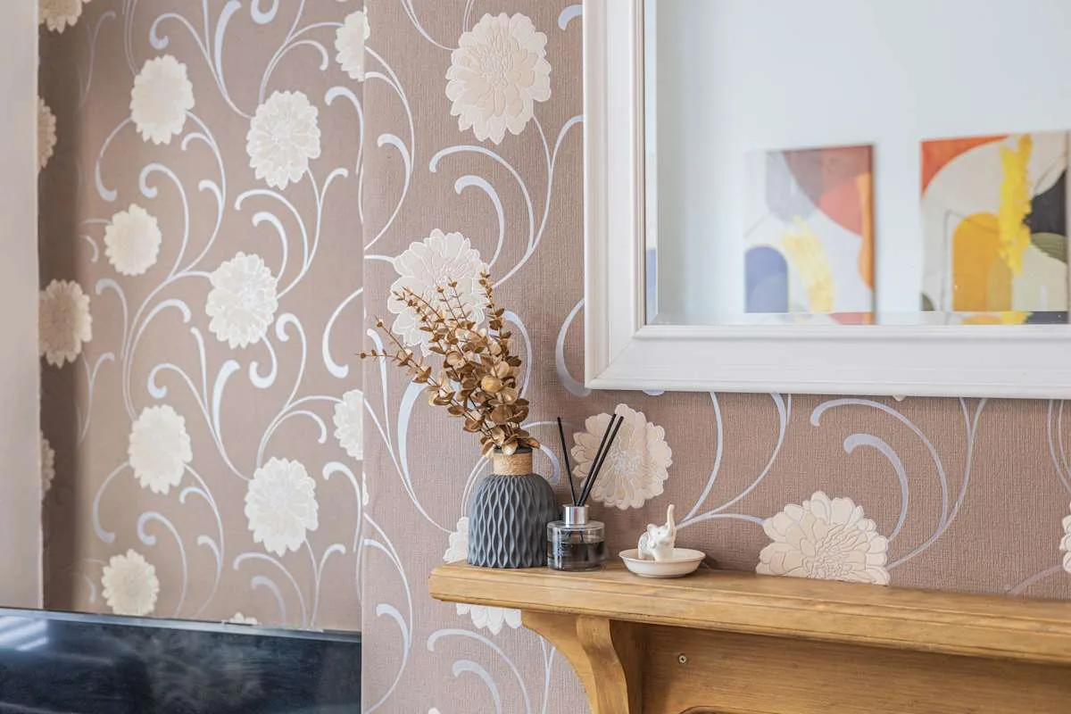 Una pared con estampado floral, una repisa de madera y cuadros.