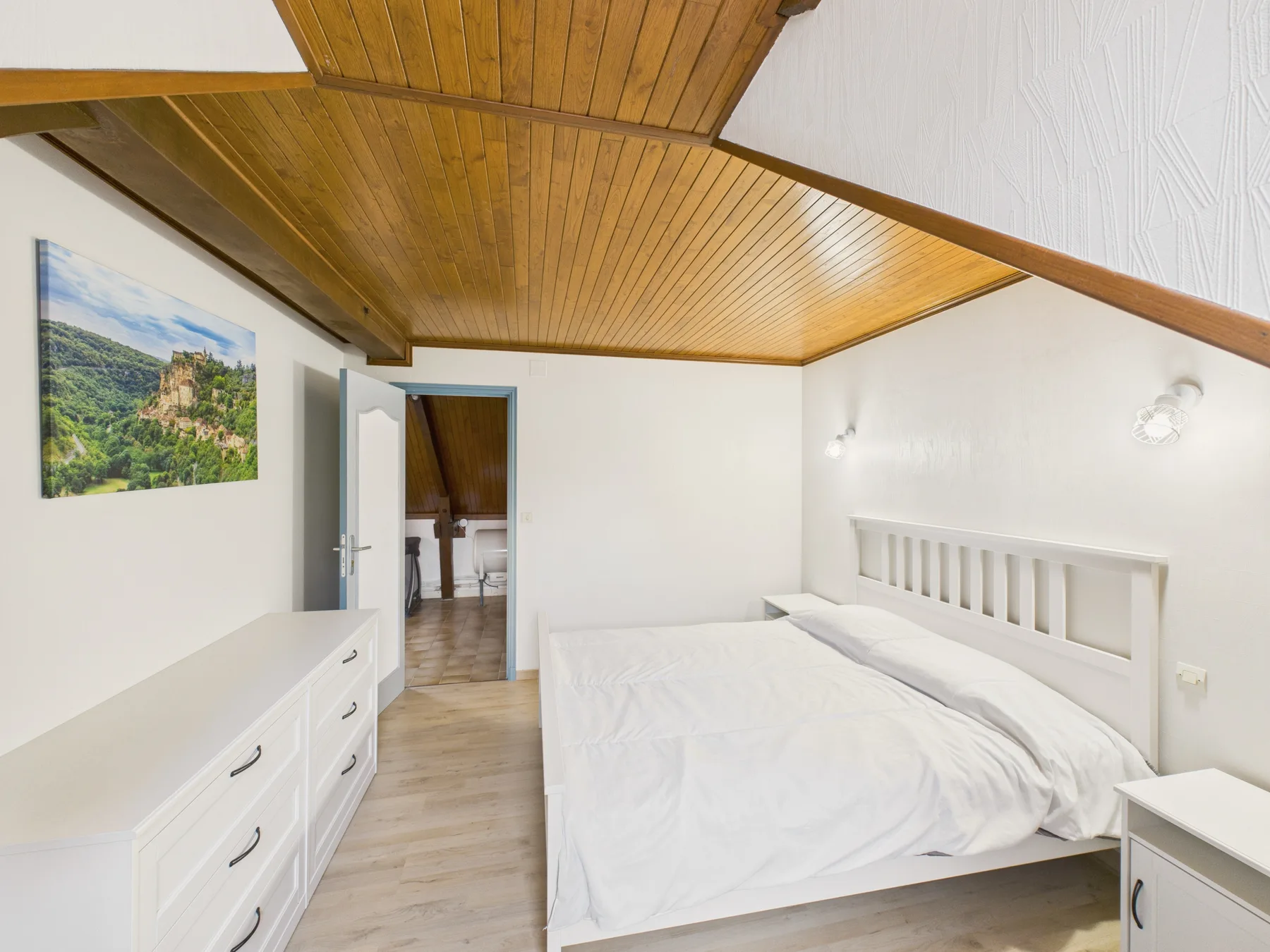Un dormitorio luminoso con una cama, muebles blancos y un techo inclinado de madera.