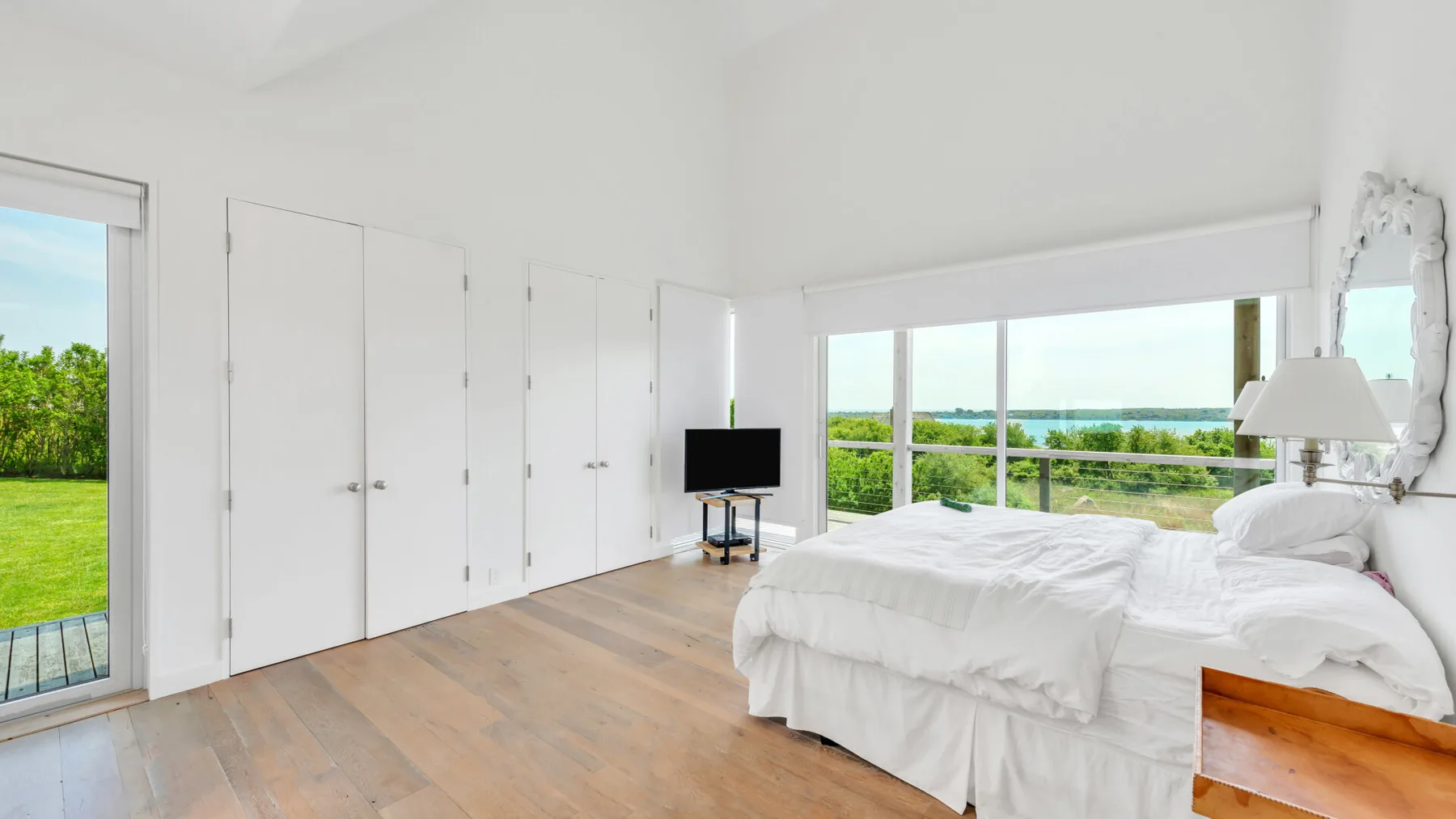 A bright bedroom with white bedding and wide windows overlooking greenery.