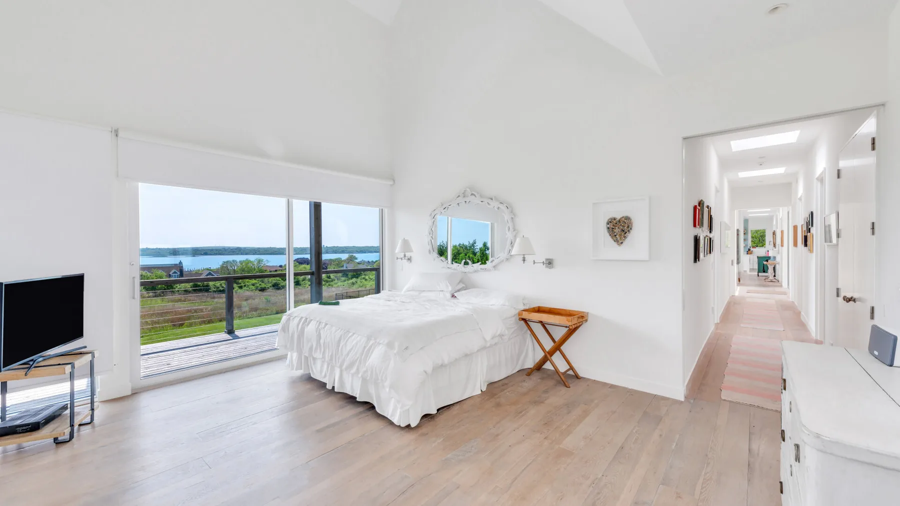 A bright bedroom with a white bed beside large windows overlooking the water.