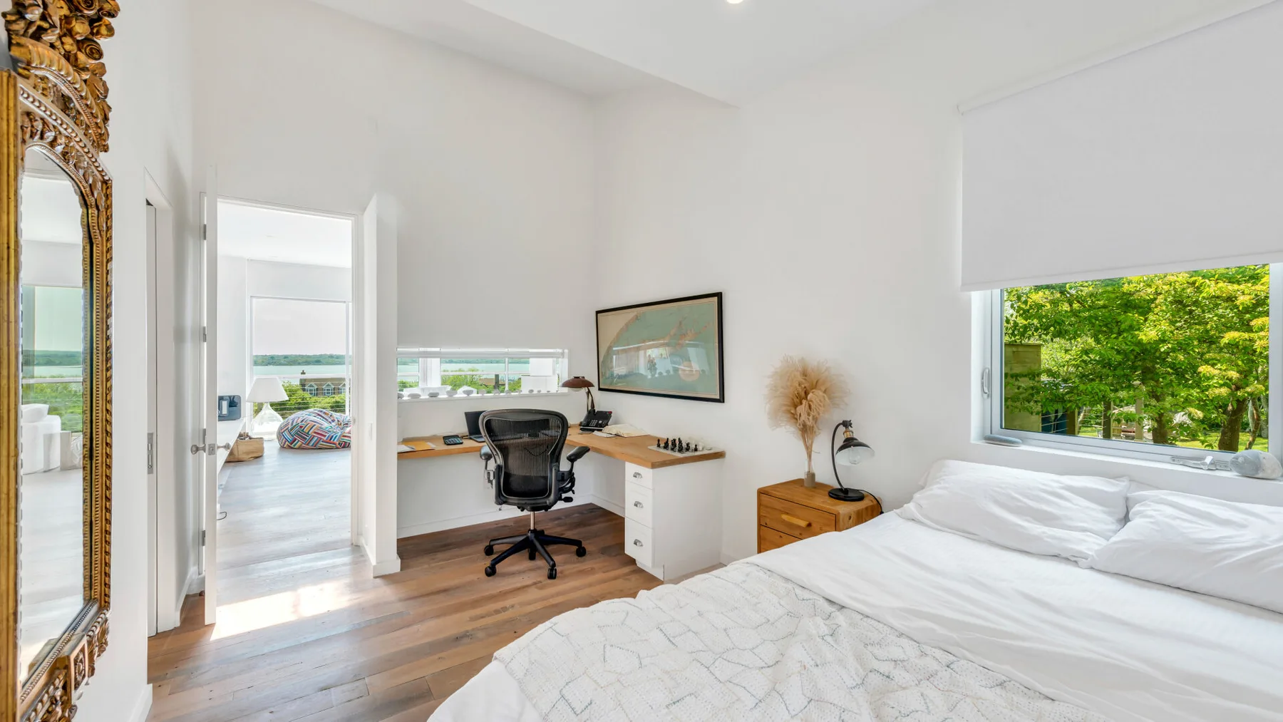 A bright bedroom with a bed, desk, and large windows overlooking greenery.