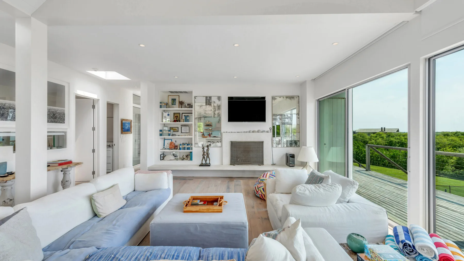 Una luminosa sala de estar con asientos blancos, un televisor y grandes ventanales hacia una terraza.