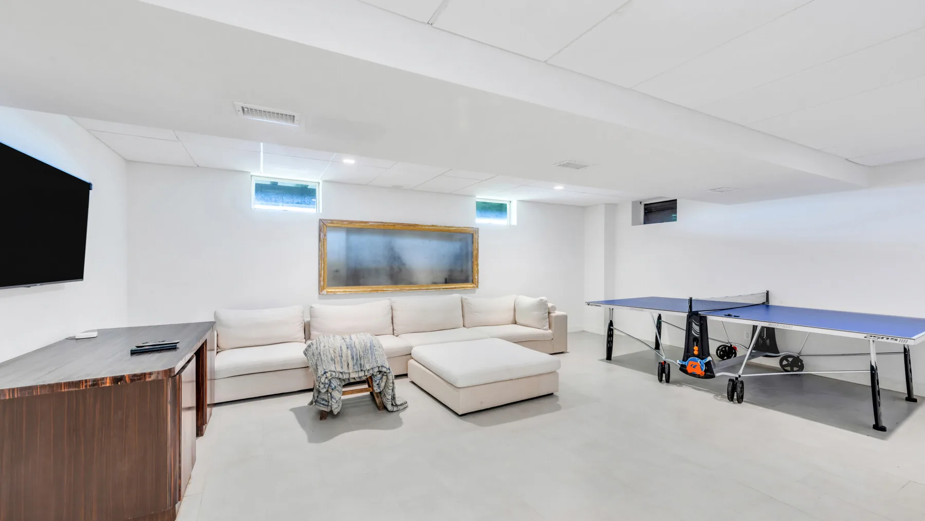 A basement recreation room has a sectional seating area and a ping-pong table.