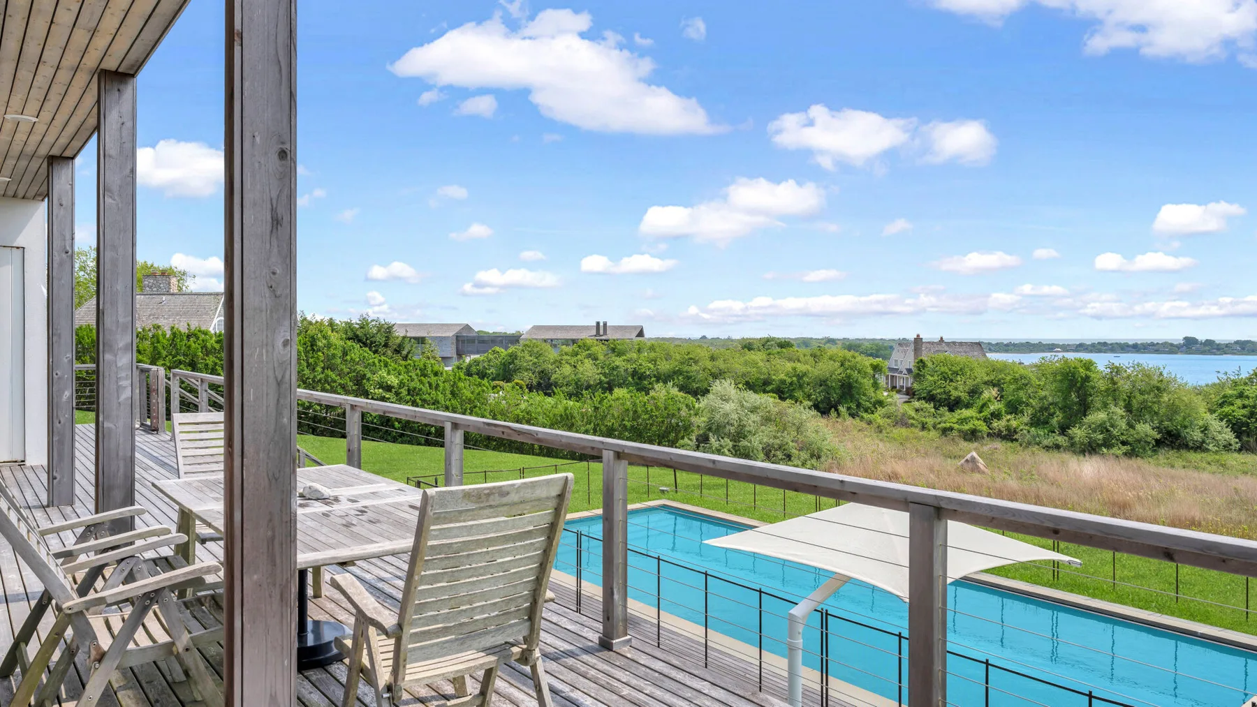 A wooden deck overlooks a swimming pool, greenery, and a stretch of water.