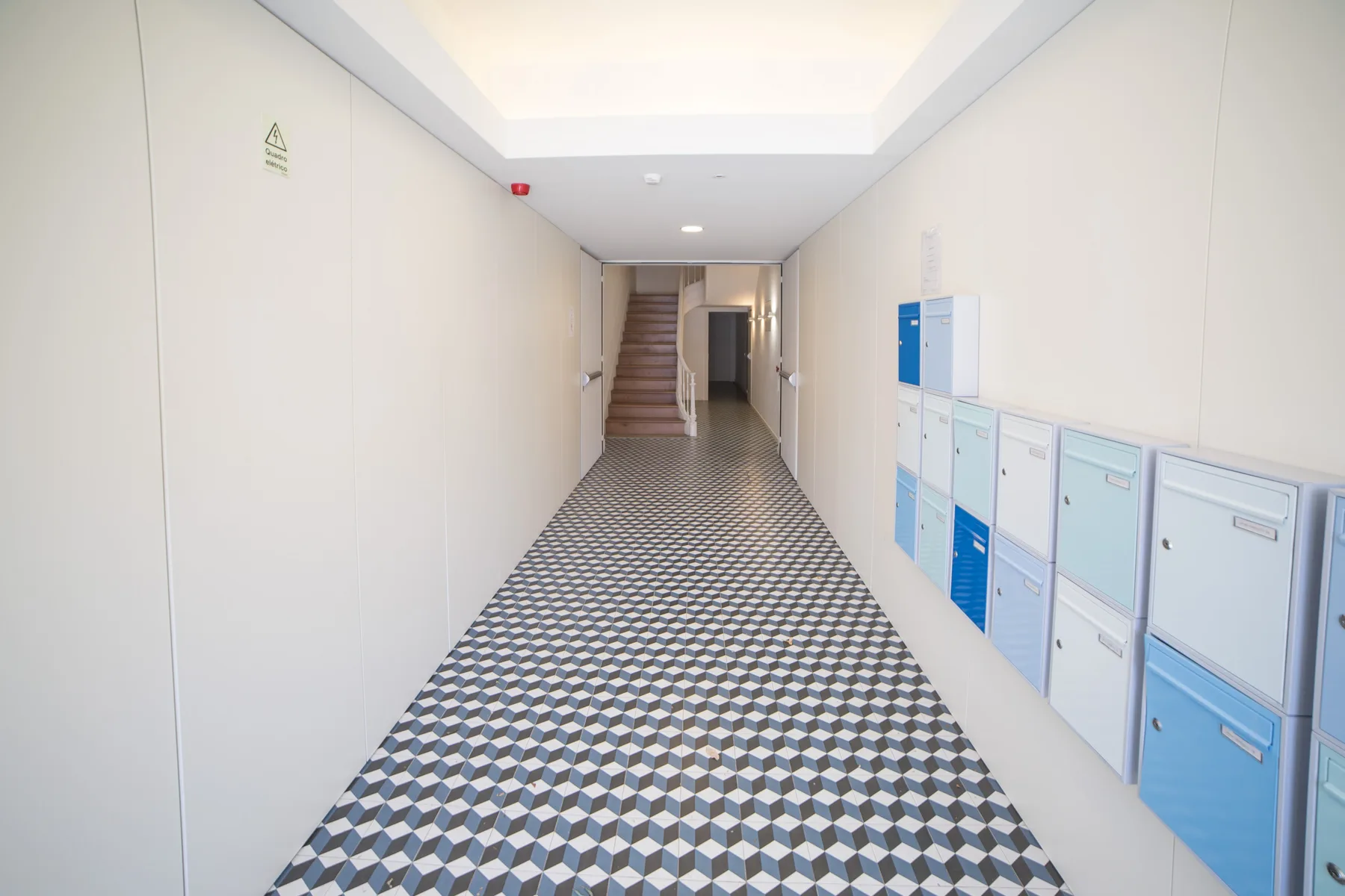 Un pasillo luminoso con suelo estampado, buzones y escaleras al fondo.