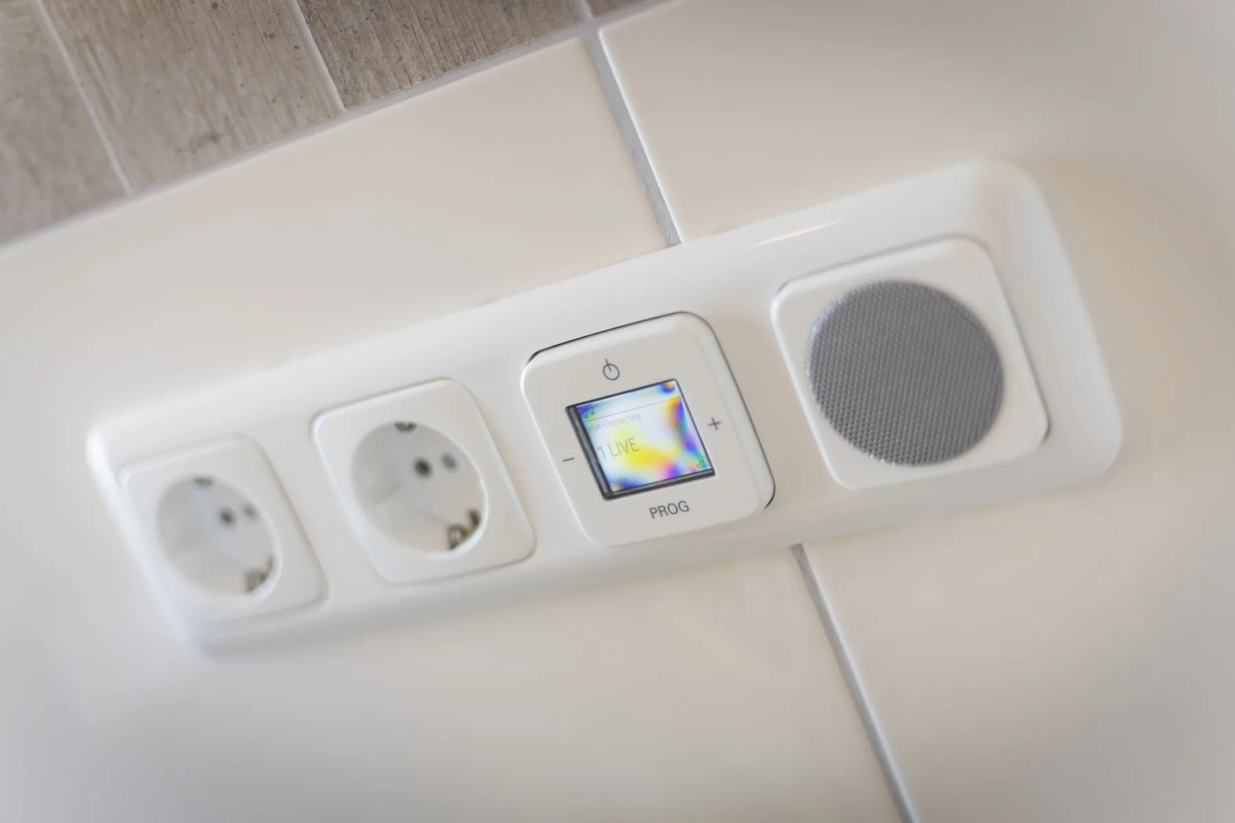 Panel en una pared alicatada con tomas de corriente, una pantalla de control y un altavoz.