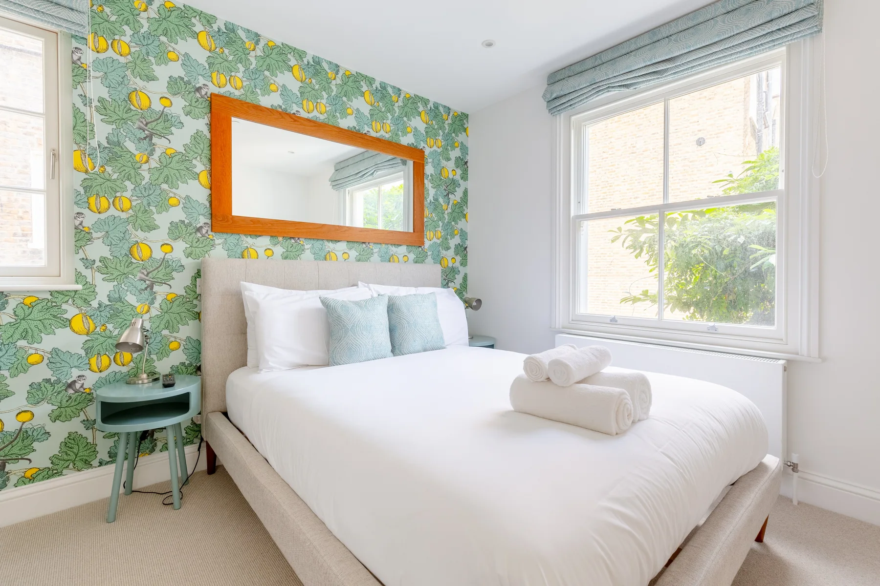 A bright bedroom with white bedding and colorful fruit-patterned wallpaper.