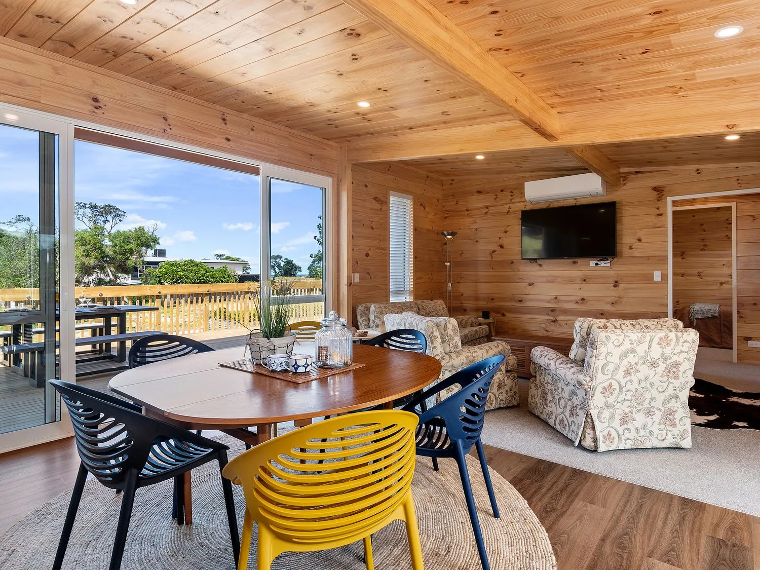 Una zona de comedor con una mesa de madera y sillas de colores junto a una sala de estar con sillones de estampado floral y un televisor montado en la pared en Matapouri Bayside Retreat, en Matapouri