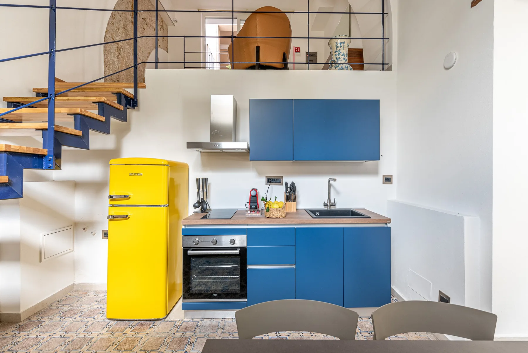 Una cocina luminosa con gabinetes azules, un refrigerador amarillo y una escalera.