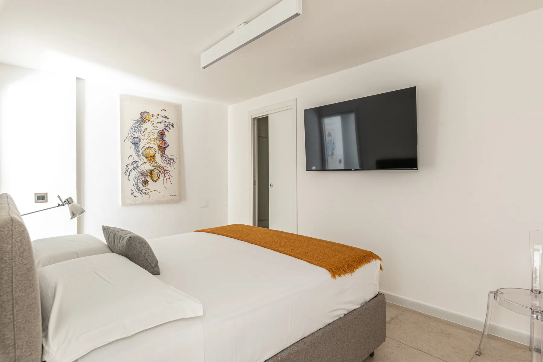 Un dormitorio luminoso con una cama, un televisor en la pared y una obra enmarcada.