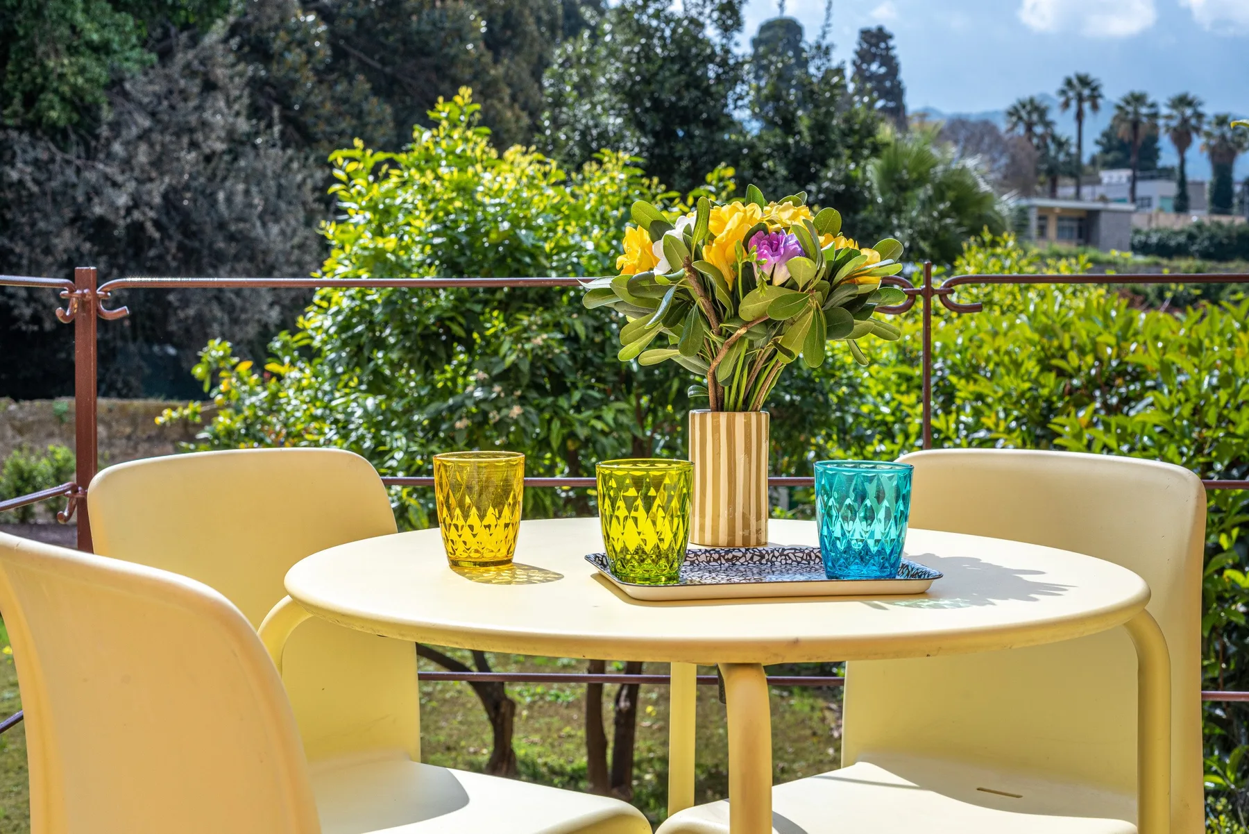 Una mesa de patio al aire libre con vasos de colores y un jarrón de flores.