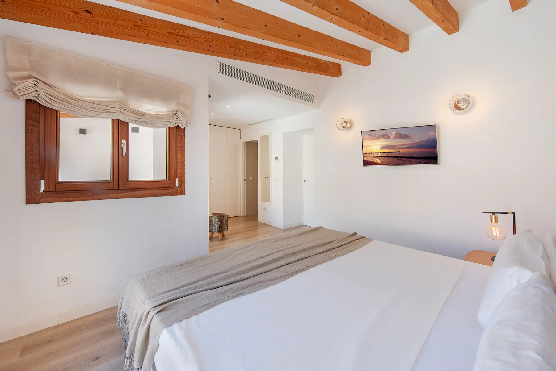A bright bedroom with a large bed beneath exposed wooden ceiling beams.
