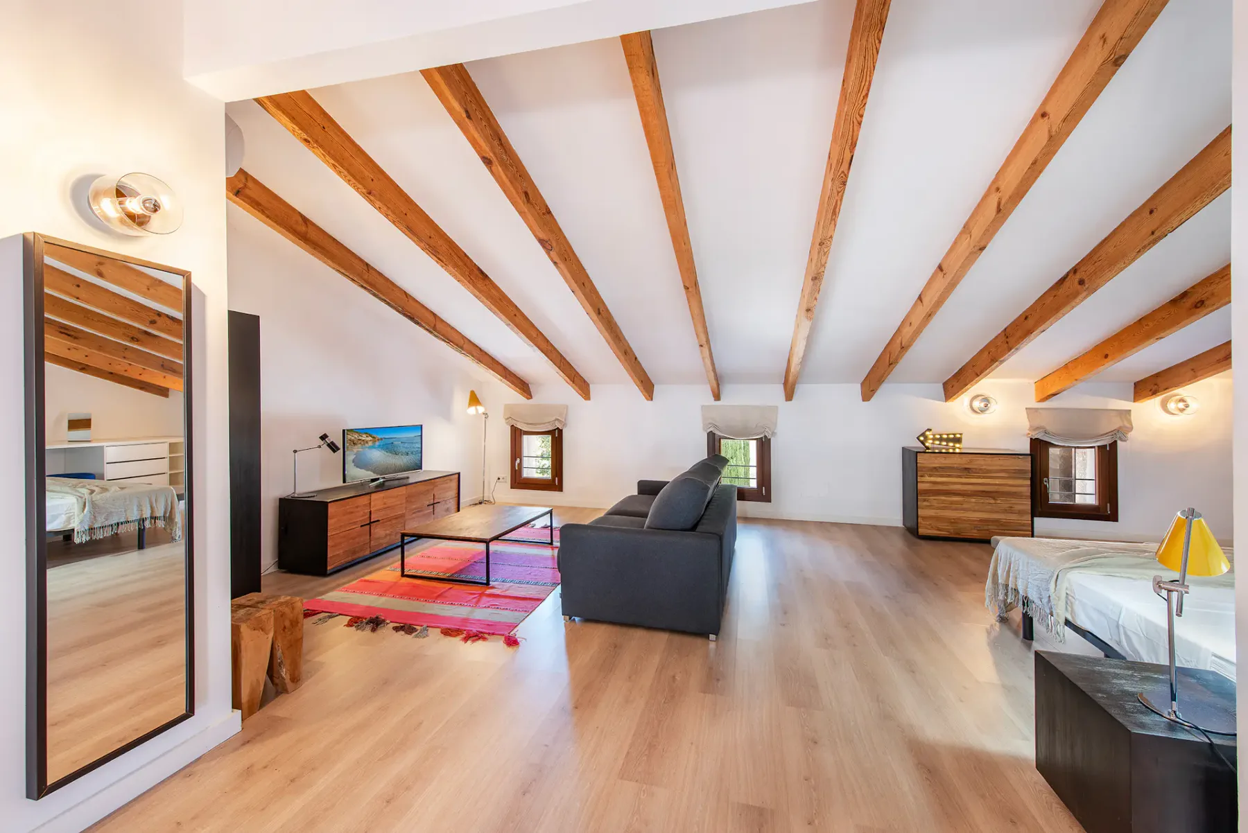A bright bedroom with exposed wooden ceiling beams, beds, and a seating area.