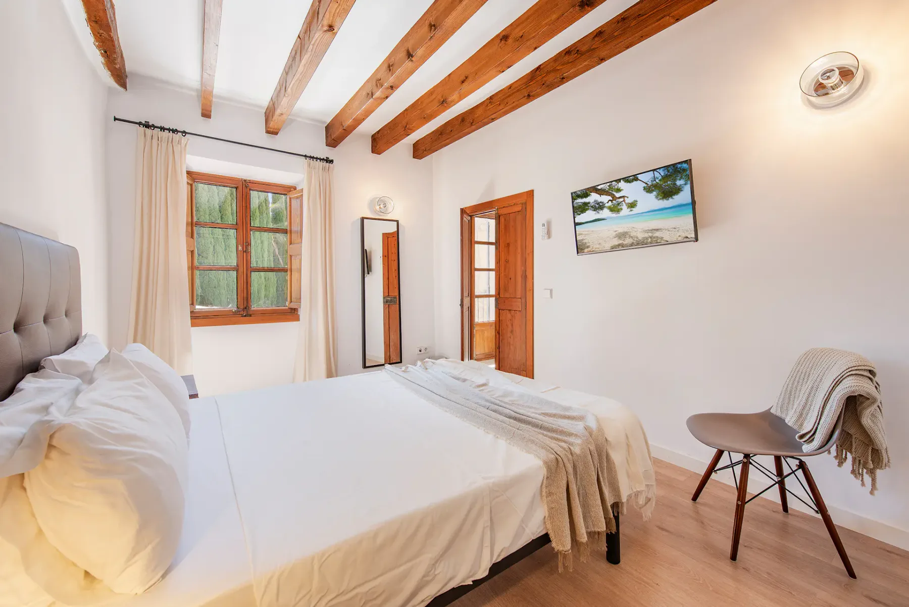 A bright bedroom with a large bed, exposed ceiling beams, and a wooden door.