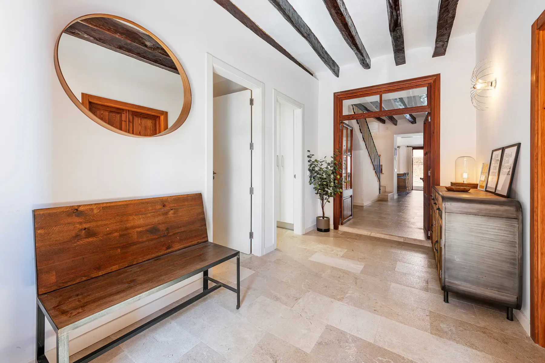 A bright entry hall with a wooden bench, oval mirror, and exposed ceiling beams.