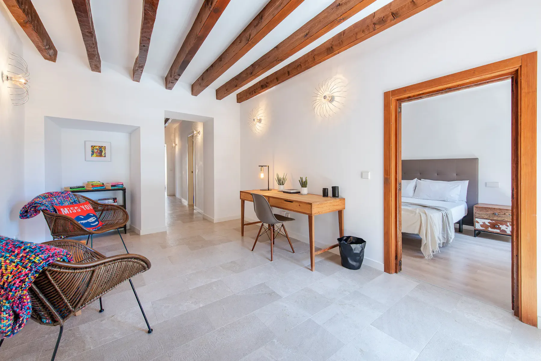 A bright room with exposed wooden ceiling beams, a desk, and seating beside a bedroom doorway.