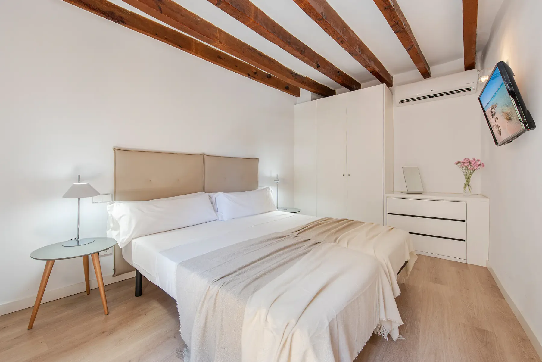 A bright bedroom with a neatly made bed and exposed wooden ceiling beams.