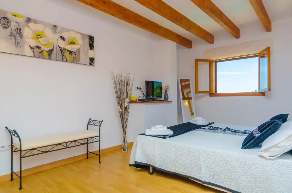 A bedroom with a neatly made bed, a wall-mounted television, and exposed ceiling beams.