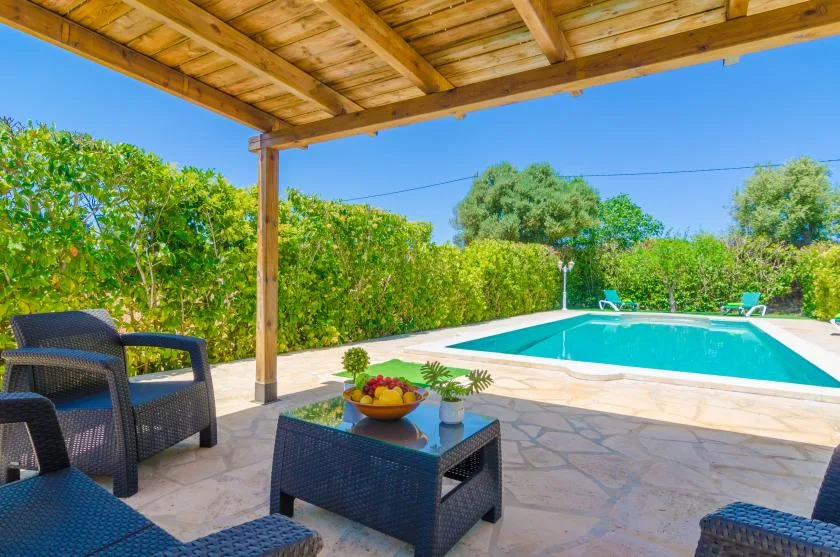 A covered patio with outdoor seating overlooks a swimming pool and green hedges.