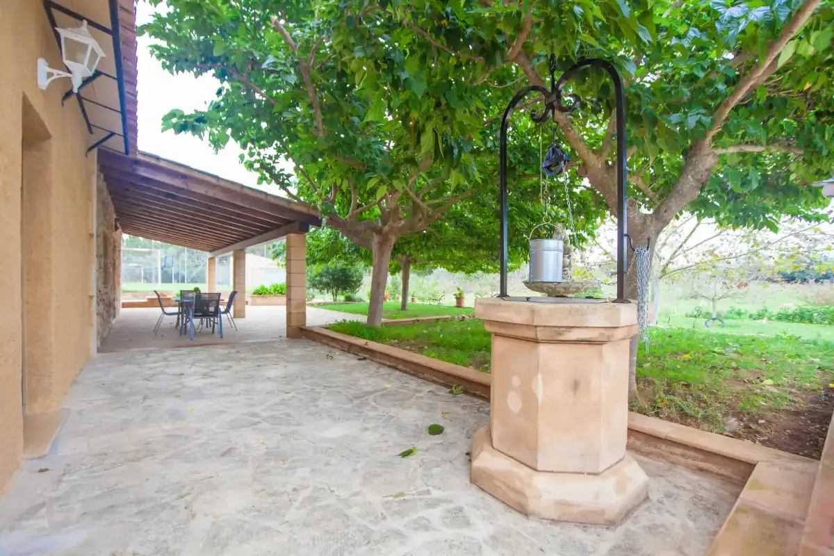 A shaded stone patio with a covered dining area and a well beneath leafy trees.