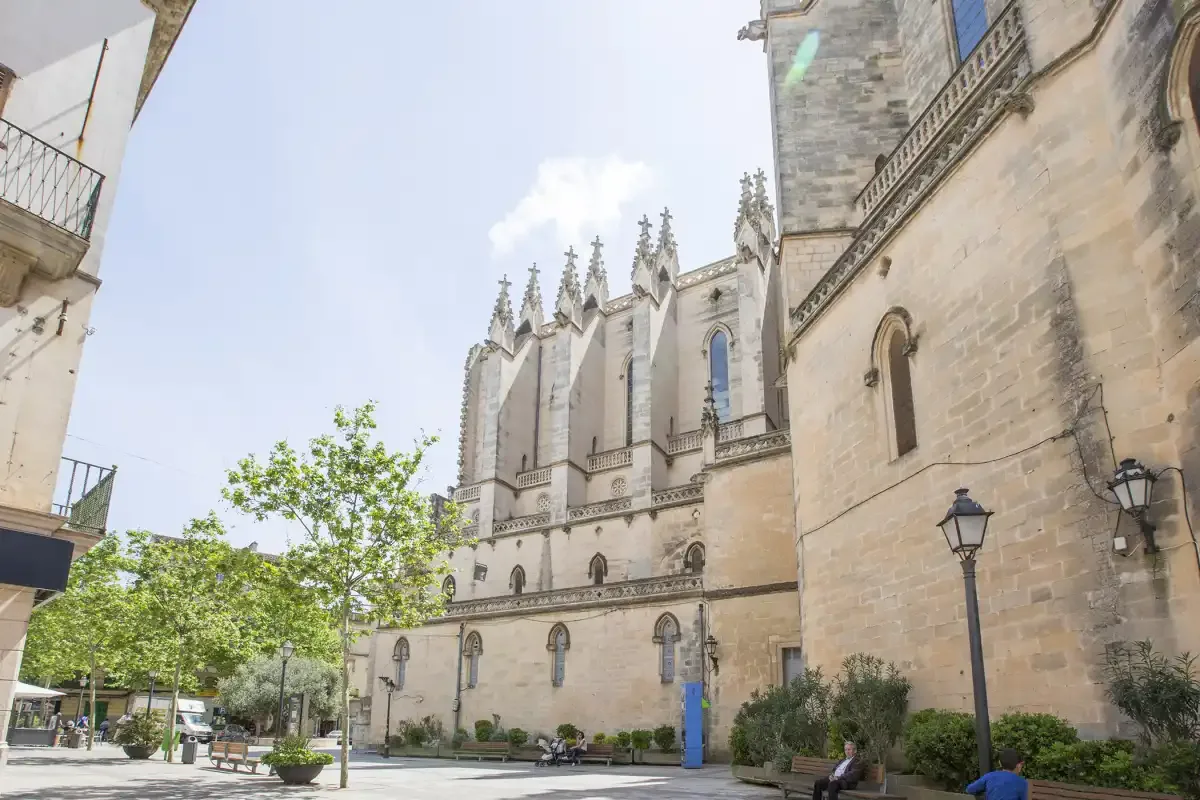 A large Gothic church stands beside a paved plaza with trees and benches.